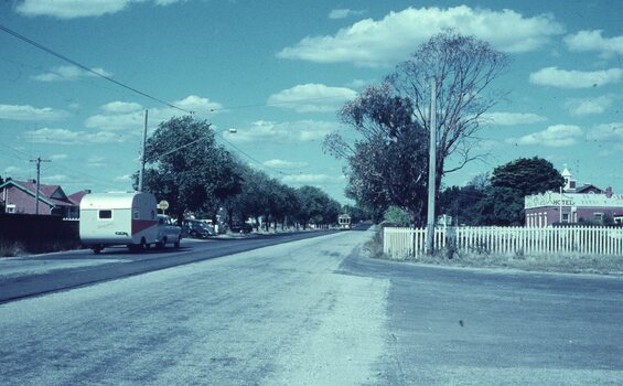 A vintage caravan is travelling on the side of a tree-lined road with a white picket fence bordering a building that appears to be a hotel with a tram at the terminus.