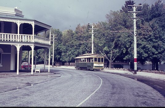 A vintage green and yellow tram is running beside a road next to a building with a white ornate balcony, surrounded by lush green trees with a lake in the background.