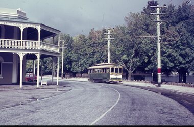 A vintage green and yellow tram is running beside a road next to a building with a white ornate balcony, surrounded by lush green trees with a lake in the background.