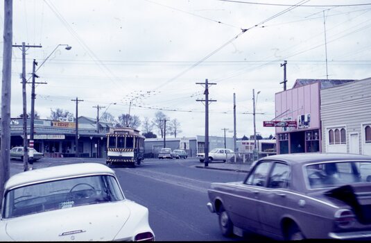 A vintage yellow and green tram travels down a street lined with shops and parked cars.