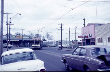 A vintage yellow and green tram travels down a street lined with shops and parked cars.
