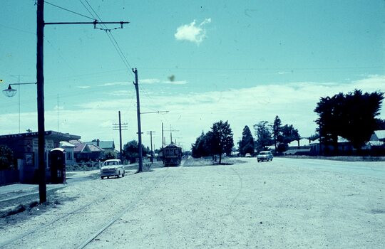 A vintage tram moves down a dirt road lined with overhead wires, with a few cars and houses visible in the background under a bright, cloudy sky.