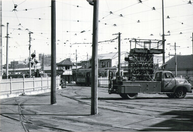 A truck with a complex overhead mechanism is parked on tram tracks near a tram station with a signal box and several trams.
