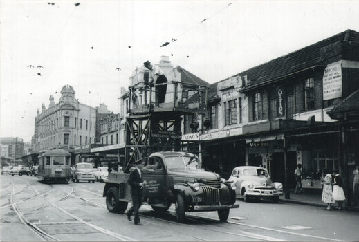 A vintage truck with "TRAMWAY EMERGENCY WAGON" emblazoned on its side is positioned on a city street with tram tracks, attending to overhead wires. A man in a hat stands on a platform attached to the truck, working on the wires, while another man walks in front of the truck. In the background, a tram, several cars, and shops with signage are visible under an overcast sky.