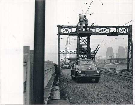 Two workers stand on a high platform attached to a lattice structure, removing overhead wires with a truck parked below on a railway track.