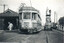 A vintage tram numbered 1838, advertising "Aspro" and "Hits the Spot", sits on tram tracks next to a tram shelter. Two individuals are working on a mobile elevated platform attached to a truck, positioned near the tram.