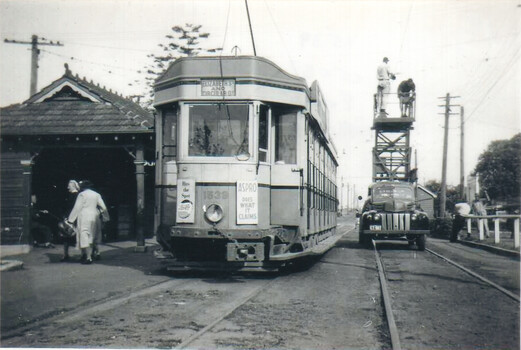 A vintage tram numbered 1838, advertising "Aspro" and "Hits the Spot", sits on tram tracks next to a tram shelter. Two individuals are working on a mobile elevated platform attached to a truck, positioned near the tram.