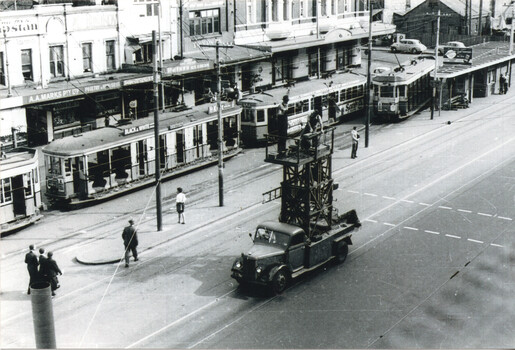 A truck with a tall, wooden tower structure attached is parked on a city street alongside several tram tracks, with trams stopped or moving in the background.