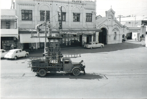 Two men on a tall platform attached to a truck are working on tramway overhead.  In the background is a sign for "The Lyric" theater, with other men and vintage cars on a street in front of shops and buildings.