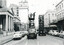 Vintage street scene showing workers on elevated platforms attached to trucks, performing overhead tramway work on a city street lined with cars and buildings.
