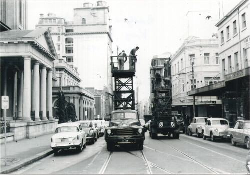 Vintage street scene showing workers on elevated platforms attached to trucks, performing overhead tramway work on a city street lined with cars and buildings.