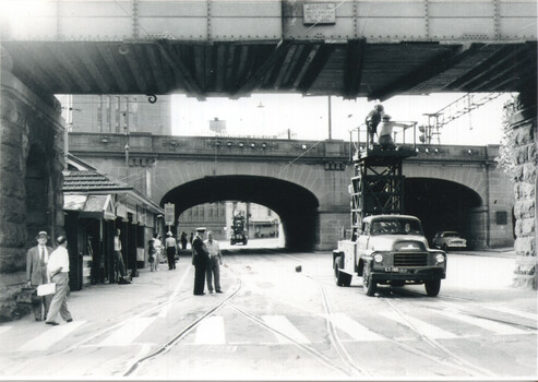 Street scene with a large railway bridge spanning overhead. A truck with a elevated platform with two workers, is positioned on the road. Several people stand on the sidewalk beside small shops.