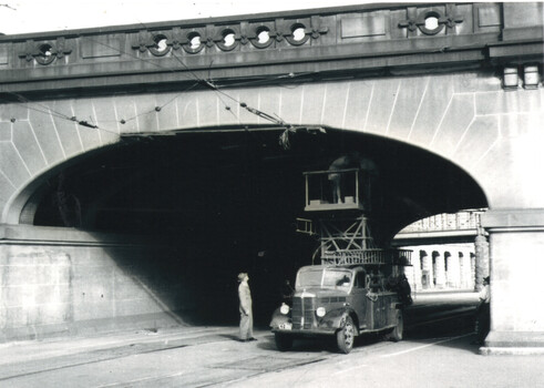 A vintage overhead tramway truck with a platform extends upwards, positioned under a stone arch bridge. A person stands near the truck, looking up.