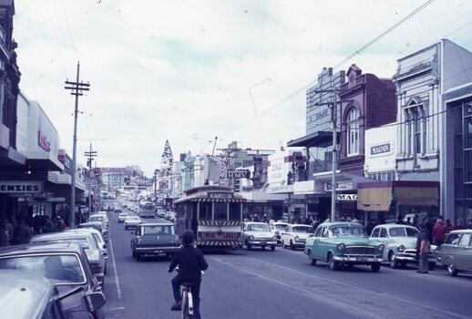 A vintage yellow tram travels down a street lined with shops and parked cars, with a person on a bicycle in the foreground.