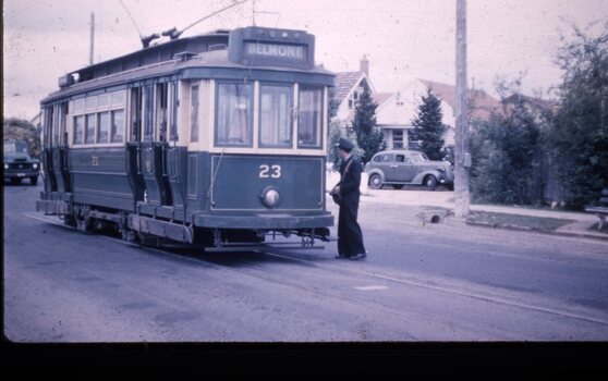 A dark green and cream vintage tram numbered 23, with "BELMONT" displayed above its front window, is stopped on a street next to a footpath. A person in uniform, the conductor, stands beside the tram's entrance, facing away from the camera. In the background, residential houses and trees are visible, and a classic car is parked in a driveway.