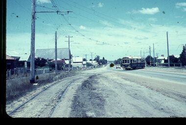 A vintage tram travels along a dusty road lined with houses and power lines.