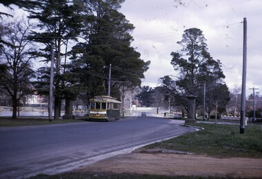 A vintage green and yellow tram is stopped at a junction on a paved road, with overhead power lines visible.