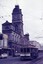 A vintage tram travels down a street in front of a grand, ornate building with a prominent clock tower, under a sky filled with overhead tram wires.