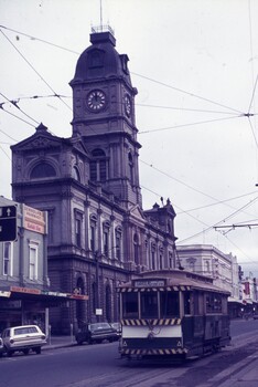 A vintage tram travels down a street in front of a grand, ornate building with a prominent clock tower, under a sky filled with overhead tram wires.