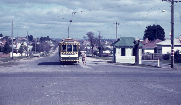A vintage tram with a white, green, and yellow livery is stopped on a street in front of a small, green-roofed shelter. A person in a striped coat stands on the street between the tram and the shelter. The street is lined with houses and trees, and the sky is cloudy.