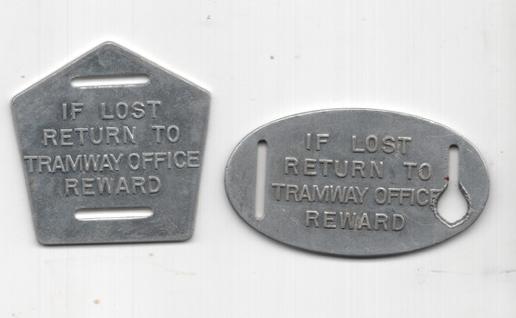 Two metallic tags, one pentagonal and one oval, with the stamped text "IF LOST RETURN TO TRAMWAY OFFICE REWARD".
