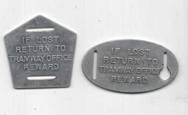 Two metallic tags, one pentagonal and one oval, with the stamped text "IF LOST RETURN TO TRAMWAY OFFICE REWARD".