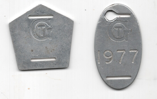 Two metal tags with a stylized "GT" logo; one pentagonal with two horizontal slots, the other oval with a cutout hole at the top and the number "1977" engraved.
