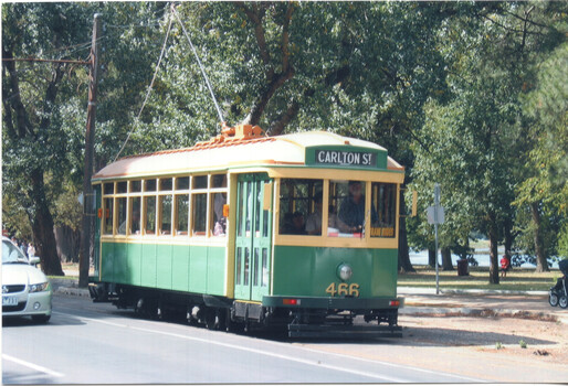 A vintage green and cream streetcar with large windows is moving along a track, with blurred trees and foliage in the background.
