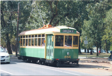 A vintage green and cream streetcar with large windows is moving along a track, with blurred trees and foliage in the background.