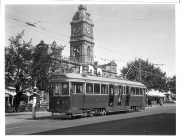 Vintage electric tram with passengers visible through windows, stopped at a street corner with a large building featuring a clock tower in the background.