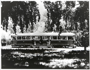 A vintage, long, double-door tram with passengers visible through the windows, stands on a track bordered by trees and foliage.