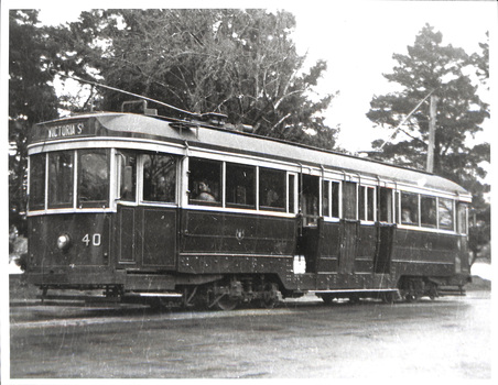 A dark, vintage tram numbered 40, with "Victoria St" displayed on its destination board, travels on tracks beside a tree-lined street.