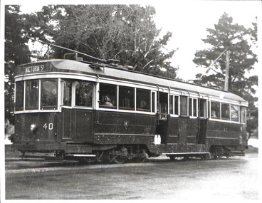 A dark, vintage tram numbered 40, with "Victoria St" displayed on its destination board, travels on tracks beside a tree-lined street.