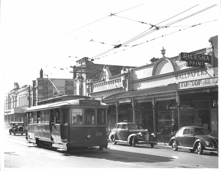 A vintage electric tram numbered 28 travels down a street lined with shops, including one with signs for "Rickshaw Paint," "Wallpapers," and "Tip Top," in front of which sit several older model cars.