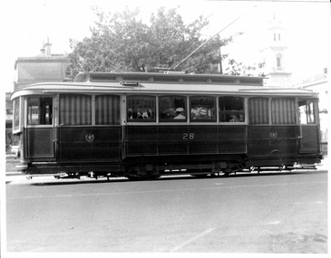 A vintage tramcar numbered 28 with visible passengers, parked on a city street with trees and a building in the background.