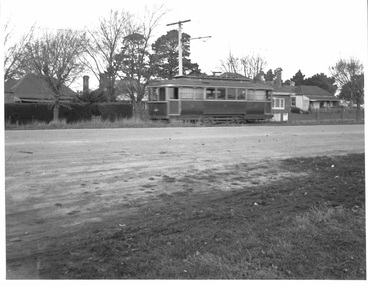A vintage wooden tramcar with multiple windows runs on tracks beside a paved road with grassy verges.