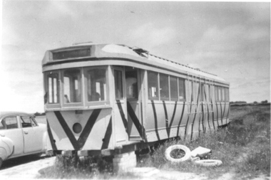 A vintage tramcar with a distinctive chevron pattern on its front, placed outdoors in a grassy area next to a classic car.