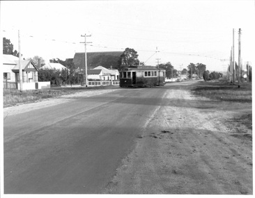 A vintage tramcar travels down a paved road in a rural setting, with buildings and utility poles lining the sides.