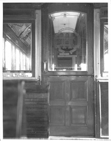 Interior of a vintage tram car showing a closed wooden door with a glass inset featuring a circular logo of the Hawthorn Tramways Trust and text. A window to the left displays a sign reading "PASSENGERS MUST NOT OBSTRUCT THE ENTRANCES."