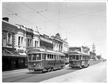 Two vintage trams with passengers travel down a street lined with historical buildings and shops.