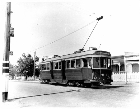 A vintage green and cream tram with an overhead power line, waits on a paved street past buildings and trees.