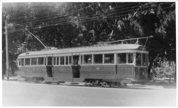A vintage tramcar with multiple windows and doors sits on tracks next to a tree-lined street.