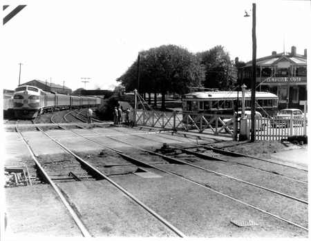 A vintage train passes through a railway crossing with a tramcar waiting nearby, a building with a sign reading "VictoriaHouse" visible in the background.