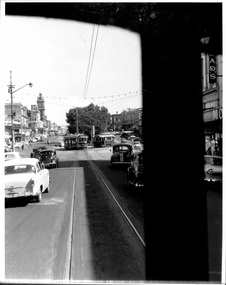 An eye-level perspective shot from a moving vehicle looking down a wide city street in the mid-20th century. Several vintage cars and at least two tramcars are visible on the road, which is lined with buildings on both sides. A partial black silhouette of a vehicle's interior frames the right side of the image.