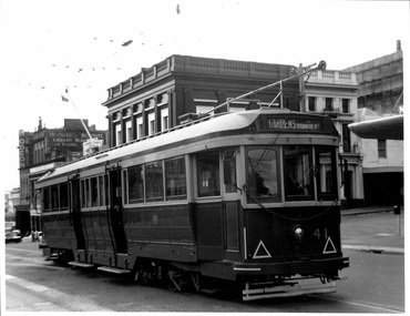 A vintage dark-colored tram with cream trim and multiple windows is stopped on a city street, with buildings visible in the background.