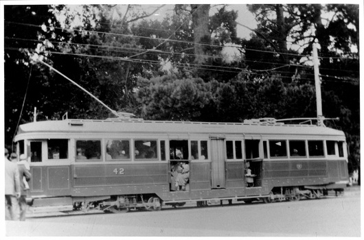 A vintage tramcar numbered 42, with passengers visible inside, travels along a track beneath overhead wires, with trees in the background.