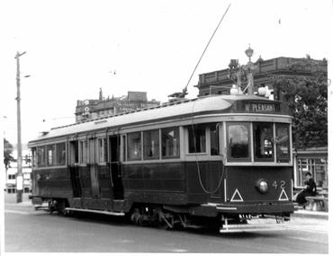 A vintage, dark-colored tram with multiple doors and windows, numbered 42, labeled "MT. PLEASANT" at the top, sits on tracks on a city street.