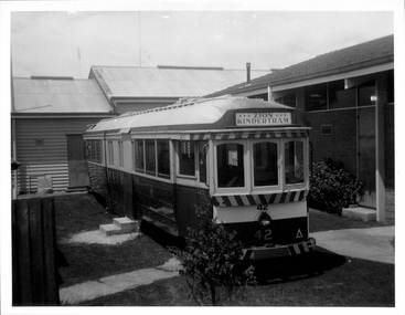 A vintage, dark-colored tramcar with white trim and the number "42" prominently displayed on the front, parked on a grassy area next to a brick building.