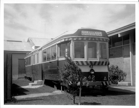 A vintage, dark-colored tramcar with yellow trim and the number "42" prominently displayed on the front, parked on a grassy area next to a brick building.