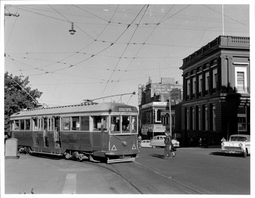 A vintage greeen and cream tram turns at a city intersection with historical buildings and cars from the mid-20th century.